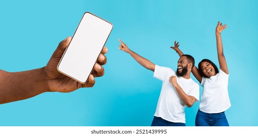 Great Offer. Cheerful African American couple dancing and pointing at giant cell phone showing blank space for mock up, celebrating win, hand holding white empty screen, blue studio background wall