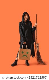 Gyönyörű fiatal boldog ázsiai nő öltözött boszorkány Halloween bevásárló táska és varázsseprű narancssárga háttérrel