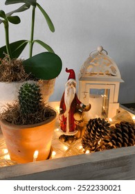 Dekorasi Natal. Nampan kayu dengan Sinterklas dan rusa kutubnya, kerucut pohon pinus kecil, lentera besi dengan lilin, vas keramik dengan tanaman dan lampu hias