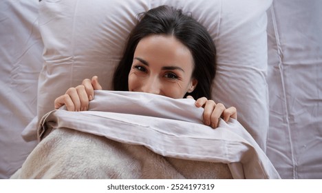Cute joyful woman with dark hair playfully hiding under soft pink sheets, peeking over the blanket with a smile. Cozy, relaxed atmosphere in a comfortable bed setting, conveying warmth and happiness