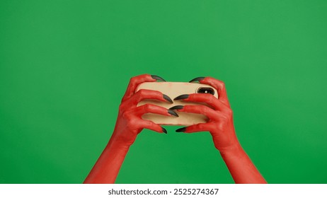 Creepy halloween devil monster hand red paint with black nails plays video game on smartphone, isolated on chroma key green screen background, workspace mockup.
