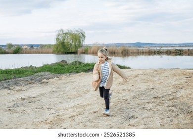 O tânără veselă aleargă de-a lungul cărării nisipoase lângă un lac liniștit, bucurându-se în aer liber în ziua înnorată.