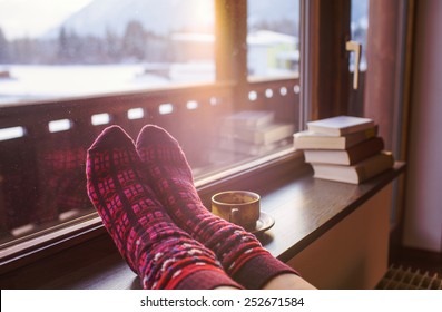 Kaki di kaus kaki wol dengan pemandangan pegunungan Alpen. Wanita bersantai dengan pemandangan gunung dengan secangkir minuman panas. Tutup dengan kaki. Konsep liburan musim dingin dan Natal.