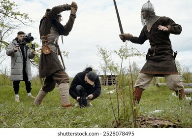 Capturando cena de reconstituição medieval com atores blindados em ação e membros da equipe filmando. Homem usando câmera profissional enquanto atores se envolvem em luta de espadas