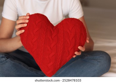 Fată care ține inimă roșie tricotată, în prim-plan. Ziua Îndrăgostiților. Pernă în formă de inimă. Cadou de Ziua Îndrăgostiților. Fată stând pe pat cu inimă de jucărie. Simbol al iubirii, cadou. Tânără romantică cu pernă