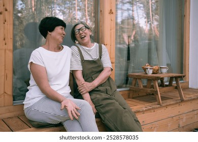 พี่สาวชาวเอเชียสูงอายุสองคนกำลังนั่งคุยกันและหัวเราะอย่างผ่อนคลายบนระเบียงบ้านในชนบทในวันฤดูร้อน ภาพระยะใกล้