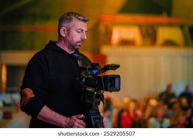 Valmiera, Lettonia - 11 ottobre 2024 - Vecpuisis. Un videografo con una felpa scura sta utilizzando una telecamera professionale su un treppiede, registrando un evento con un pubblico sullo sfondo, fuori fuoco.