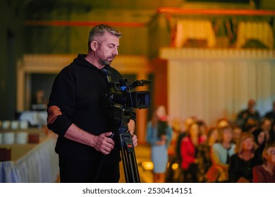 Valmiera, Lettonia - 11 ottobre 2024 - Vecpuisis. Un videografo con una felpa scura sta utilizzando una telecamera professionale su un treppiede, registrando un evento con un pubblico sullo sfondo, fuori fuoco.