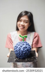 صورة لامرأة مبتسمة تحمل كعكة عيد ميلاد أرجوانية تشبه "Gomu Gomu No Mi" في سلسلة الأنمي One Piece. ضوء طبيعي، خلفية بيضاء.