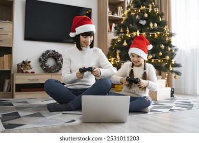 Caucasian mother and daughter wearing Santa hats play video games with joysticks on laptop near Christmas tree. Room filled with festive decorations creates joyful holiday atmosphere.