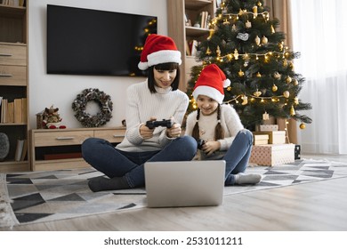 Caucasian mother and daughter wearing Santa hats play video games using joysticks and laptop. Christmas tree decorates living room creating festive atmosphere.