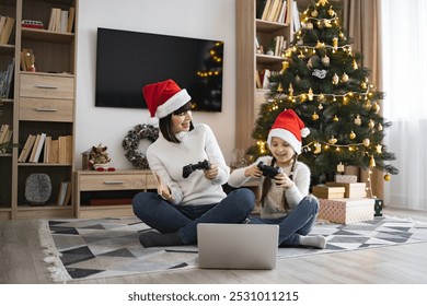 Caucasian mother and daughter wear Santa hats while playing video games using joysticks and laptop in festive room. Christmas tree decorated with lights and ornaments adds warmth to cheerful family