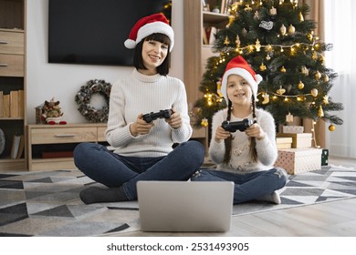 Smiling Caucasian mother and daughter enjoy playing video games together with joysticks on laptop. Cozy living room decorated for Christmas with tree and gifts.