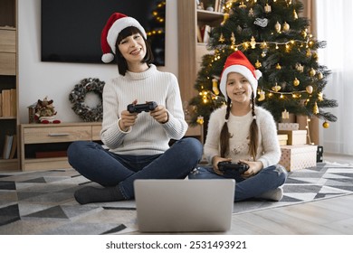 Caucasian mother and young daughter enjoy playing video games together during Christmas. Both wear Santa hats, sitting on floor by Christmas tree with lights and ornaments.