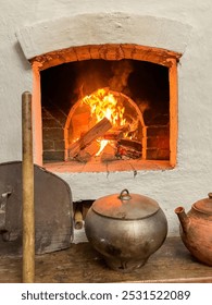 Un horno de ladrillos con fuego encendido en su interior. Una cuchara de madera está apoyada contra el horno. Una tetera está sobre una mesa al lado del horno.