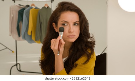 Concentrated serious Caucasian woman female girl blogger vlogger looking mirror reflection. Model streamer preparing for shooting blog video applying blush powder cosmetics face makeup brush in studio
