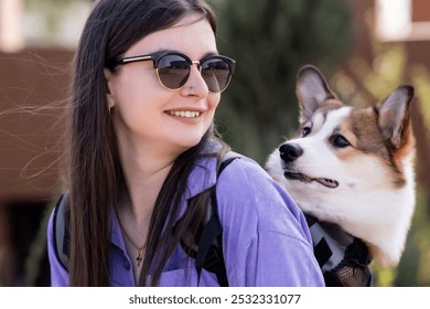Ritratto di una bella ragazza sorridente con occhiali da sole con un cucciolo di Pembroke Welsh Corgi in uno zaino speciale sulla schiena. Passeggiata in una giornata di sole in un parco cittadino. Cagnolino felice. Concetto di cura, vita animale