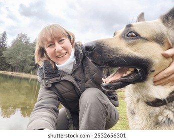 Fille adulte avec un chien de berger prenant un selfie près de l'eau d'un lac ou d'une rivière en montagne. Femme d'âge moyen et gros animal de compagnie dans la nature. Amitié, amour et plaisir
