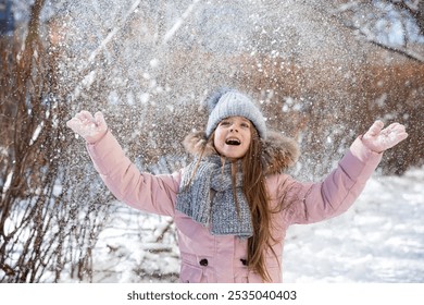Winter lifestyle portret van vrolijk meisje in gebreide muts die sneeuw in de lucht gooit. Kind lacht en heeft plezier in het sneeuwpark. Sneeuwvlokken vallen naar beneden. Vrijheid en geluk concept.