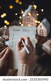 Rédiger à la main les résolutions du Nouvel An 2025 en décembre, le soir du Nouvel An. Plan, objectifs, idées, projets. Ambiance de vacances à la maison, lumières de Noël, décoration festive, biscuits au pain d'épices faits maison