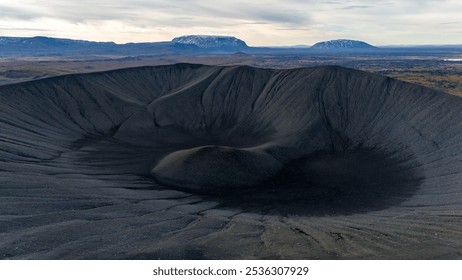 Pemandangan udara dari kawah gunung berapi yang besar dan terjal, dikelilingi oleh lanskap tandus di Islandia, dengan gunung-gunung di kejauhan di bawah langit mendung yang redup, menyoroti kekuatan alam yang dahsyat.