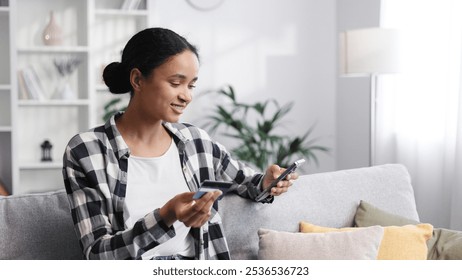 Smiling pretty young African American brunette girl holding cell phone and credit card while sitting on the sofa. Happy woman shopping online using mobile payment. Electronic banking application.