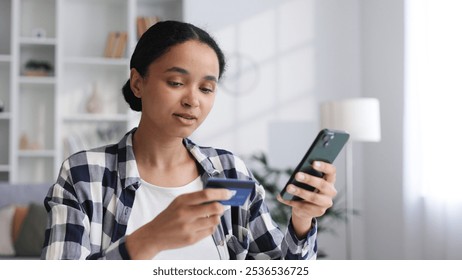 Smiling pretty young African American brunette girl holding cell phone and credit card while sitting at table. Happy woman shopping online using mobile payment. Electronic banking application.