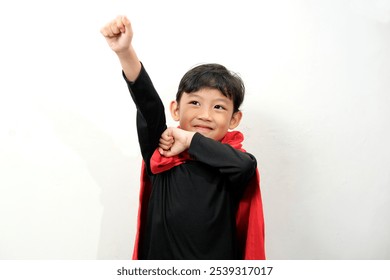 一个小男孩打扮成超级英雄，披着红色斗篷，高兴地举起一只手臂，摆出英雄的姿势。非常适合想象、自信、童年梦想和顽皮活力的概念。