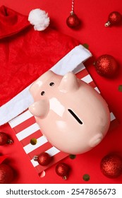 Celengan di kotak hadiah, bola dan topi Santa di latar belakang merah, tampak atas