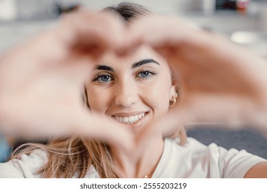 สาวสวยทำท่าชูมือรูปหัวใจพร้อมนิ้วที่พับไว้ ยิ้มแย้มแจ่มใส มองกล้อง ถ่ายภาพบุคคล หญิงสาวผู้มีเสน่ห์และมองโลกในแง่ดี แสดงให้เห็นถึงสัญลักษณ์แห่งความรัก รู้สึกมีความสุขขณะโพสท่าอยู่คนเดียวที่บ้าน