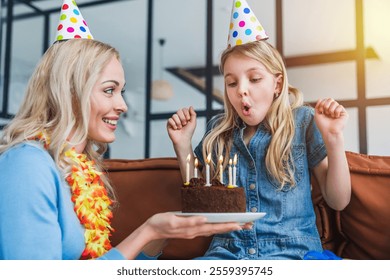 Une adorable petite fille souffle les bougies de son gâteau d'anniversaire à la maison. Une jeune et belle maman et sa petite fille, coiffée d'un chapeau, célèbrent l'anniversaire de leur fille avec un gâteau et des bougies à la maison.