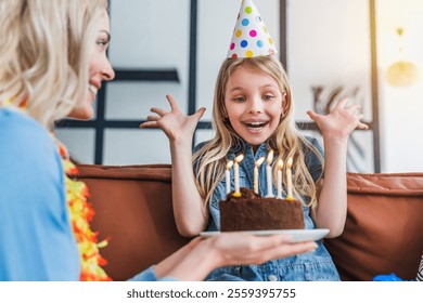 Photo d'une mère souriante et de sa petite fille surprise, coiffées de chapeaux de fête, célébrant l'anniversaire de leur fille avec un gâteau. La famille félicite la petite fille à la maison, avec un gâteau et des bougies.