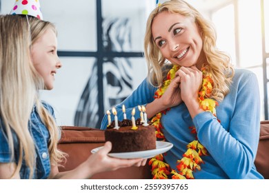 Gros plan sur une petite fille présentant un gâteau d'anniversaire à sa maman à la maison. Une jeune et belle maman et sa petite fille, coiffée d'un chapeau, célèbrent l'anniversaire de leur enfant avec un gâteau et des bougies dans le salon.