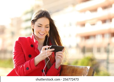 Woman watching videos in a smart phone with earphones sitting in a park with an unfocused buildings in the background