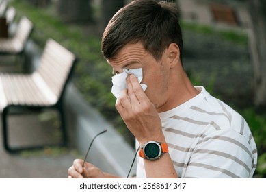 Một chàng trai trẻ đang lau nước mắt bằng khăn giấy trong khi cầm kính ở công viên, cảm thấy khó chịu do dị ứng theo mùa và tác động của phấn hoa trong không khí.