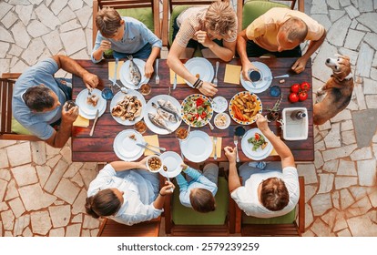 Vue plongeante d'une famille joyeuse réunie autour d'une table à manger extérieure, savourant un repas méditerranéen composé de poisson frais, de légumes et de salades, des plats colorés sur une terrasse rustique, ambiance estivale, convivialité.