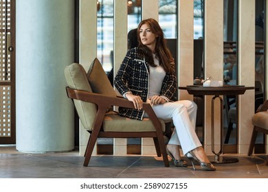 A well-dressed woman sits gracefully on a plush chair in a modern hotel lounge, relishing her tranquil surroundings. Soft lighting enhances the cozy ambiance while she appears lost in thought.