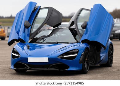O mașină sport albastru strălucitor, cu uși distinctive care se deschid în sus, este expusă pe asfalt într-o zonă deschisă într-o zi însorită. Designul său elegant și culorile vibrante sporesc puterea vehiculului.