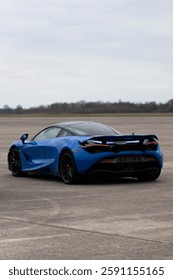 O mașină sport albastră vibrantă stă parcată pe o pistă pustie, evidențiindu-și caracteristicile aerodinamice și designul modern sub un cer înnorat