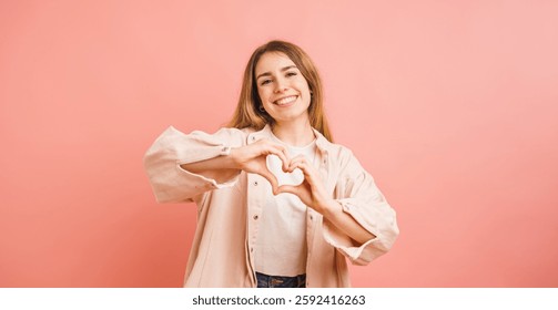 Giovane donna sorridente che disegna un cuore con le mani su sfondo rosa