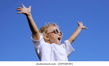 Blond haired girl wearing white shirt and sunglasses radiating pure childhood joy, stretching arms skyward against vibrant blue background, embodying carefree enthusiasm and youthful energy