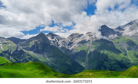 Un magnifique paysage de montagnes enneigées dominant des collines verdoyantes. Le ciel est d'un bleu profond parsemé de nuages ​​blancs et duveteux. Un décor idéal pour toute aventure en plein air.