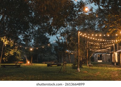 Ein gemütliches Lagerfeuer im Freien, umgeben von rustikalen Holzbänken und Lichterketten, schafft eine friedliche Abendatmosphäre – perfekt für Glamping unter dem Sternenhimmel in einem naturnahen Waldrefugium.