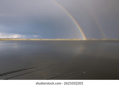 Varas de pesca em um píer de madeira sobre um lago calmo com um arco-íris duplo no céu, natureza tranquila após a chuva.
