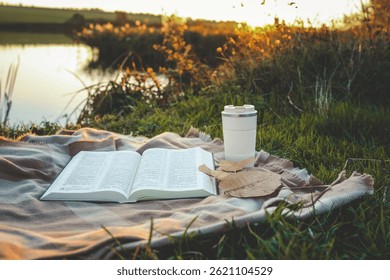 Bible on plaid blanket with coffee cup and autumn leaves near peaceful riverbank at sunset. Tranquil outdoor scene of spiritual reflection, nature, and calm during golden hour.