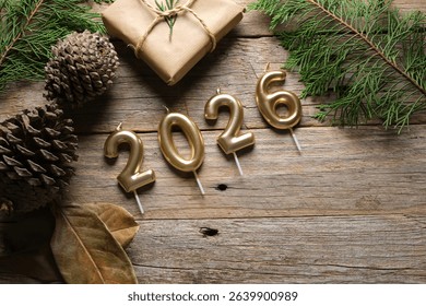 Des bougies dorées représentent 2026, entourées de pommes de pin et de verdure sur une surface en bois rustique, rehaussant l'ambiance festive des célébrations du Nouvel An.