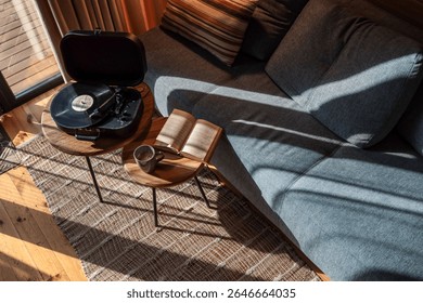 Vista de arriba hacia abajo de un acogedor rincón de lectura y música en una cabaña iluminada por el sol, con un libro abierto, una taza de café y un reproductor de discos de vinilo en mesas auxiliares de madera cerca de un sofá gris.