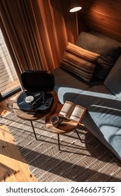 Vista de arriba hacia abajo de un acogedor rincón de lectura y música en una cabaña iluminada por el sol, con un libro abierto, una taza de café y un reproductor de discos de vinilo en mesas auxiliares de madera cerca de un sofá gris.