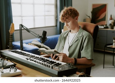 Adolescent caucazian stând la birou citind o carte în timp ce înregistrează muzică cu microfon și tastatură digitală într-un studio modern de acasă, cu chitară electrică vizibilă în fundal