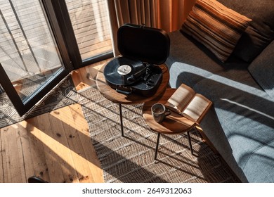 Vista de arriba hacia abajo de un acogedor rincón de lectura y música en una cabaña iluminada por el sol, con un libro abierto, una taza de café y un reproductor de discos de vinilo en mesas auxiliares de madera cerca de un sofá gris.